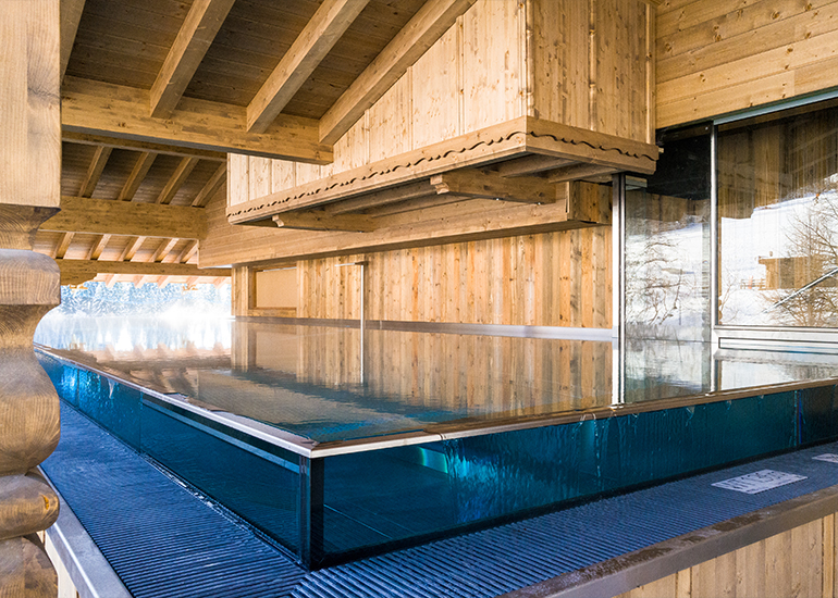 Ein moderner Außenpool mit einem großen Holzüberhang. Das Wasser ist klar und spiegelt die umliegende Architektur wider. Der Bereich ist von warmen Holzpaneelen umgeben, was eine gemütliche Atmosphäre schafft.